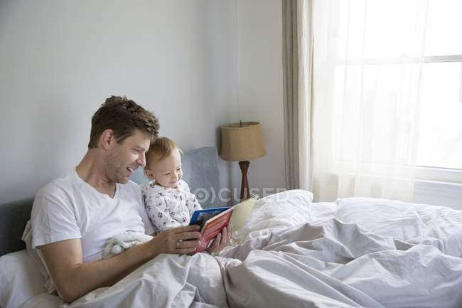Padre e hijo pequeño, sentados en la cama, leyendo el libro juntos - foto de stock