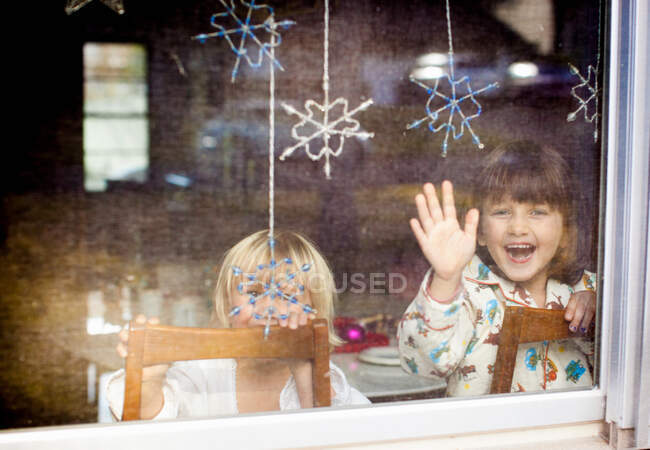 Дети смеются и машут в окно с висящими снежинками — стоковое фото