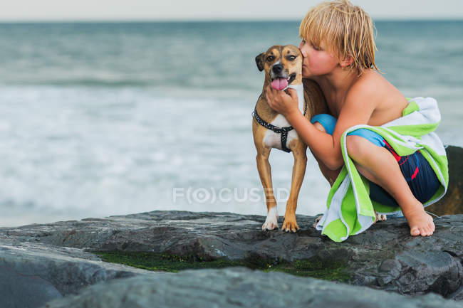 Niño agachado en las rocas en la playa, abrazando perro mascota - foto de stock