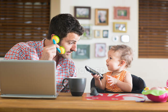 Мужчина и ребенок сидят за кухонным столом, играя со смартфоном и игрушечным телефоном — стоковое фото