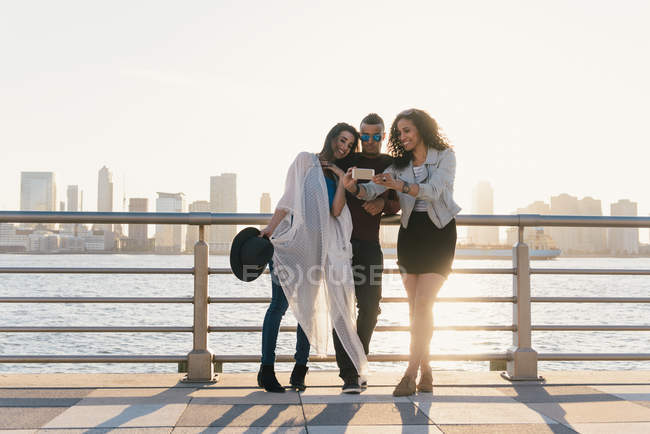 Tres amigos adultos medios tomando selfie de teléfonos inteligentes en el paseo marítimo, Nueva York, EE.UU. - foto de stock