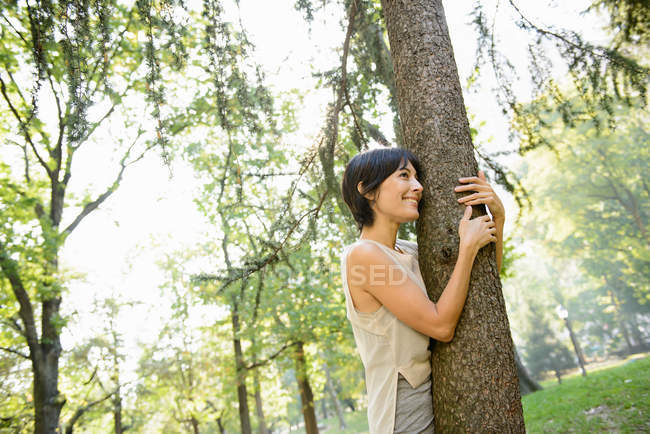 Улыбающаяся женщина обнимает дерево в парке — стоковое фото