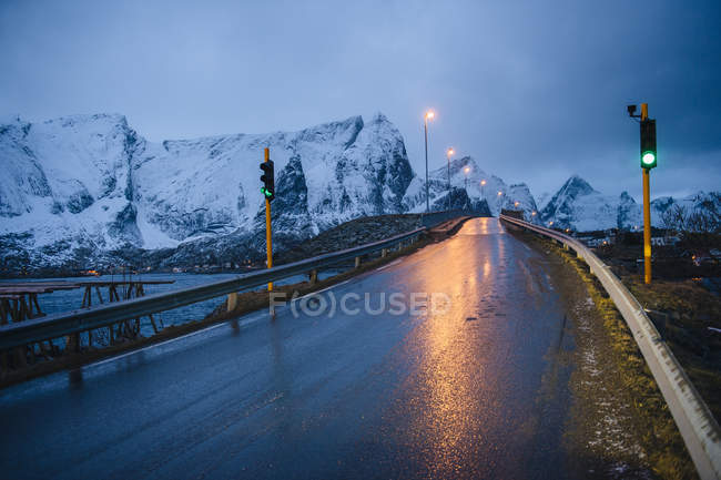 Estrada molhada com reflexo de luzes de rua e montanhas cobertas de neve — Fotografia de Stock
