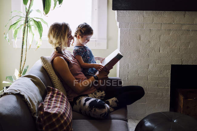 Mulher no sofá com a filha da criança lendo livro de histórias — Fotografia de Stock