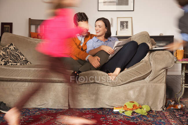 Взрослая пара отдыхает на диване с детьми, двигающимися вокруг них — стоковое фото