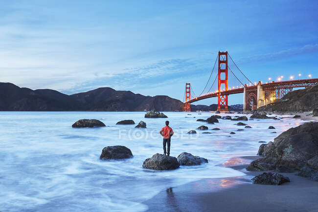 Szenische Ansicht der Golden Gate Bridge bei Sonnenuntergang mit einer Person, die Marshall 's Beach im Vordergrund steht. San Francisco, Kalifornien, USA — Stockfoto
