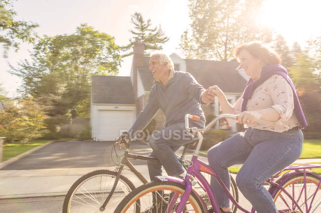 Romantic senior couple holding hands cycling along suburban road — Stock Photo
