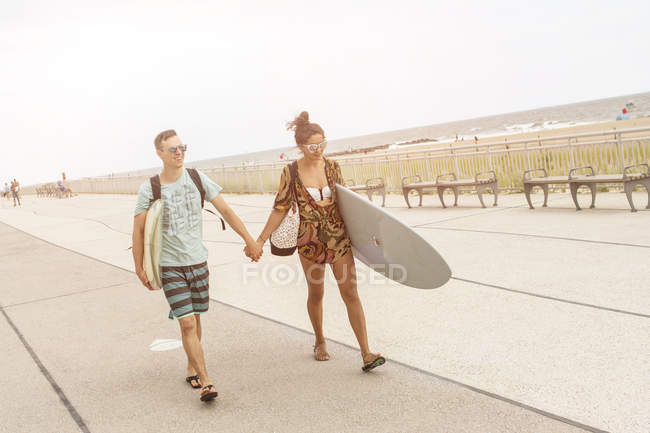 Junges Paar hält Händchen und trägt Surfbretter am Rockaway Beach, New York State, Vereinigte Staaten — Stockfoto