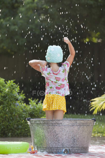 Вид сзади на девушку, стоящую в пенной ванне в саду, плескающую мыльные пузыри — стоковое фото