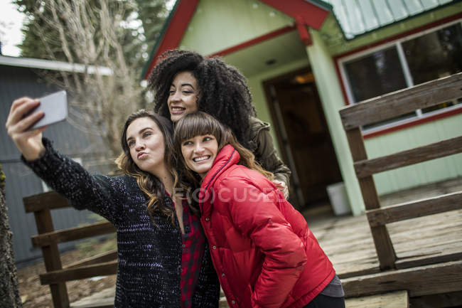 Three friends standing beside cabin, taking selfie, using smartphone — Stock Photo