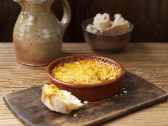 Bowl of smoked cheese bake, crusty bread on wooden chopping board — Stock Photo