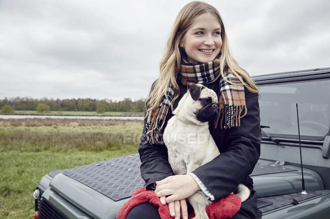 Jeune femme assise sur un véhicule hors route dans un champ avec chien sur les genoux — Photo de stock