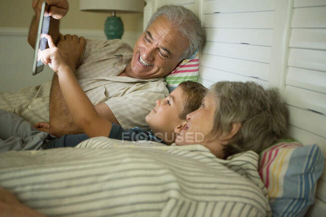 Abuelos acostados con su nieto y mirando la tableta digital - foto de stock