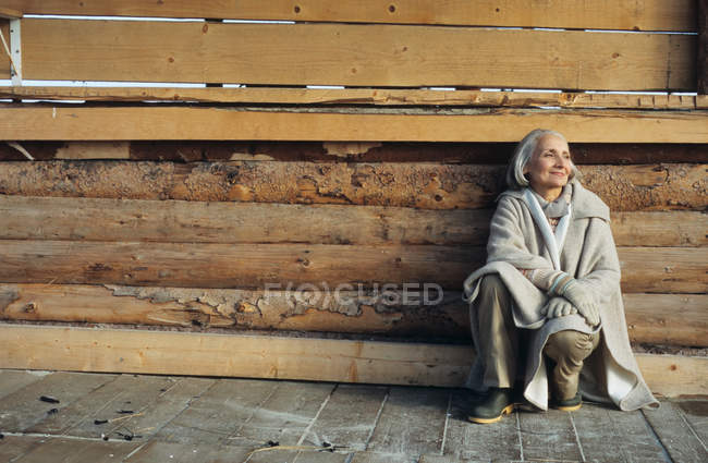 Donna anziana accovacciata vicino a casa di legno — Foto stock
