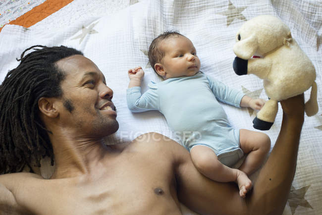 Padre e hijo acostados en la cama jugando con un juguete suave - foto de stock