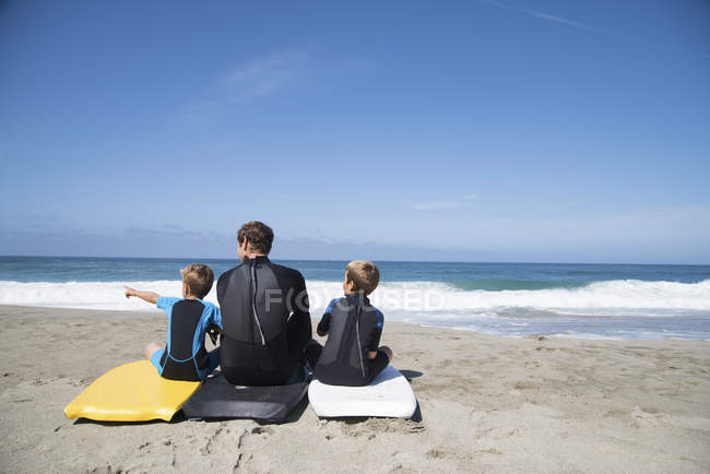 Vista trasera del hombre y dos hijos sentados en bodyboards señalando, Laguna Beach, California, EE.UU. - foto de stock