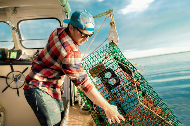 Молодой человек поднимает клетку с омарами с лебедки на рыбацком судне на побережье штата Мэн, США — стоковое фото