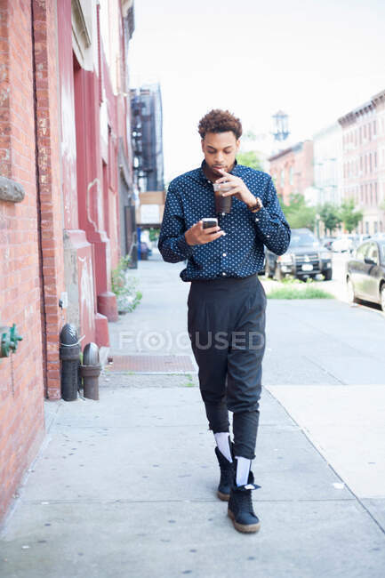 Молодой человек идет по улице с сотовым телефоном и напитками — стоковое фото