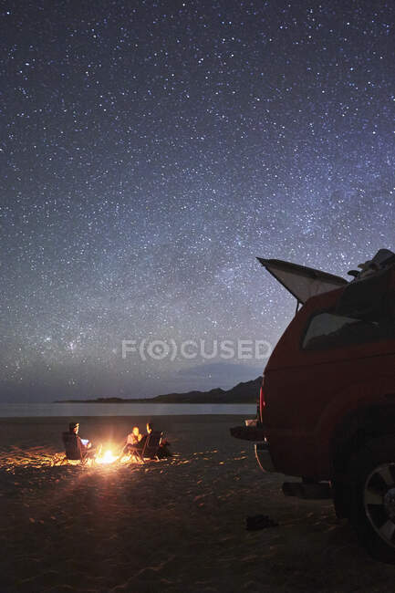Amici campeggi auto si riuniscono intorno a un falò sulla spiaggia sotto un cielo notturno pieno di stelle. — Foto stock