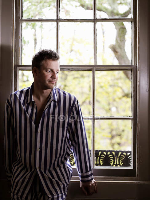 Hombre sentado junto a la ventana en pijama - foto de stock