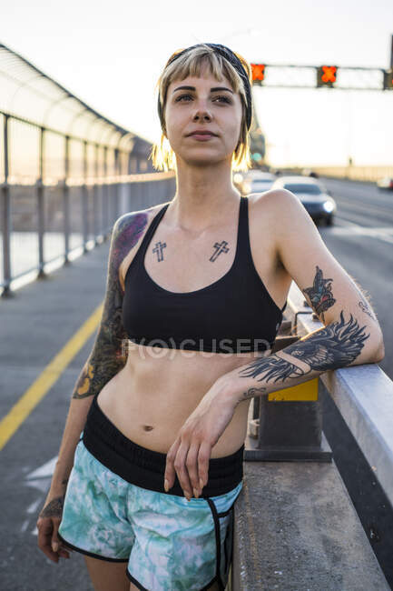 Joven mujer tatuada corriendo en el puente tomando un descanso con la puesta de sol detrás - foto de stock
