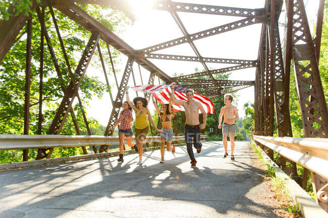 Grupo de amigos corriendo en el puente sosteniendo bandera americana - foto de stock