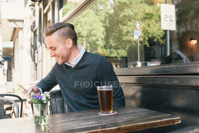 Jeune homme assis à l'extérieur du bar, en utilisant smartphone — Photo de stock