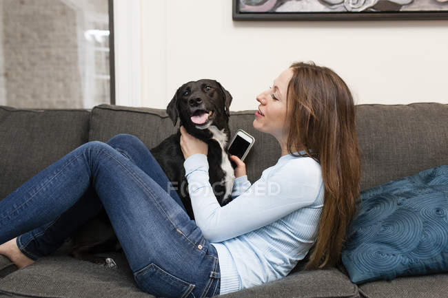 Вид сбоку молодой женщины на диване, держащей смартфон и играющей с собакой — стоковое фото