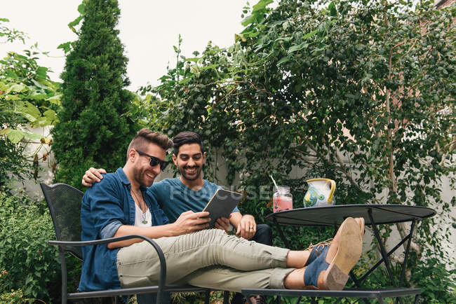 Young male couple sitting in garden and looking at digital tablet — Stock Photo