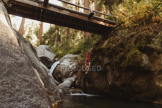 Junger Mann sitzt in Hängematte, aufgehängt von Brücke, Mineral King, Sequoia National Park, Kalifornien, USA — Stockfoto