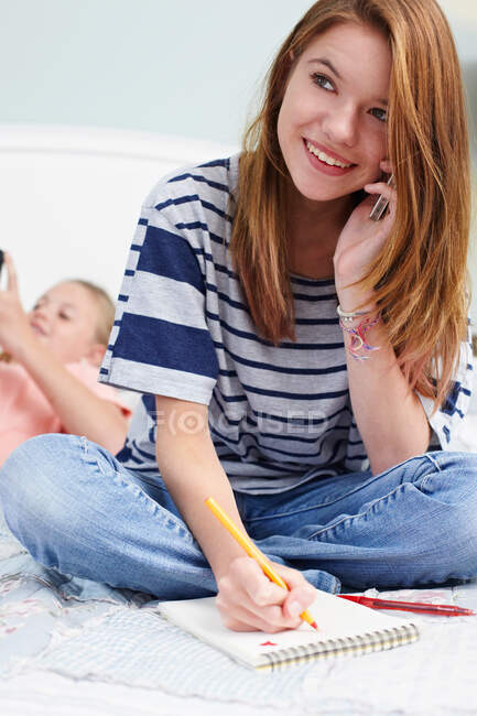 Teenage girl sitting on bed with notepad, using cellphone — Stock Photo