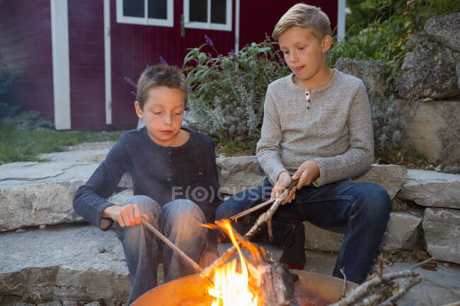 Два мальчика с палками сидят у костра в саду на закате — стоковое фото