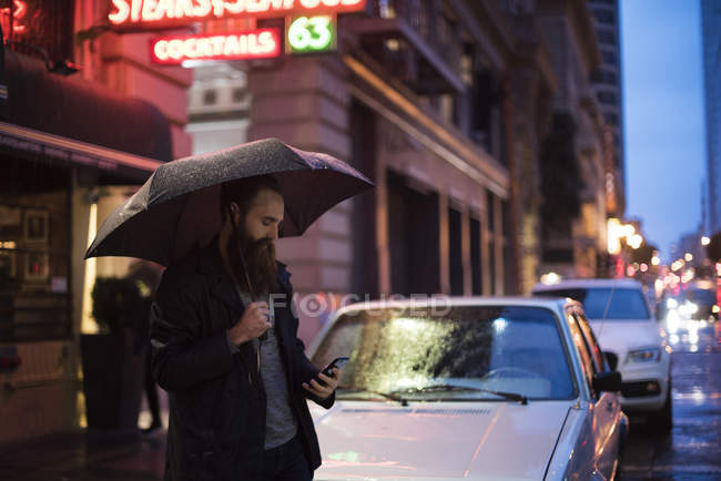Людина ходить по місту вночі, використовуючи парасольку, дивлячись на смартфон, центр міста, Сан-Франциско, Каліфорнія, США — стокове фото