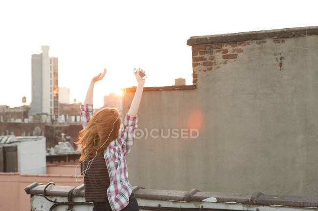 Молода жінка слухає музику зі зброєю, піднятою на даху міста — стокове фото