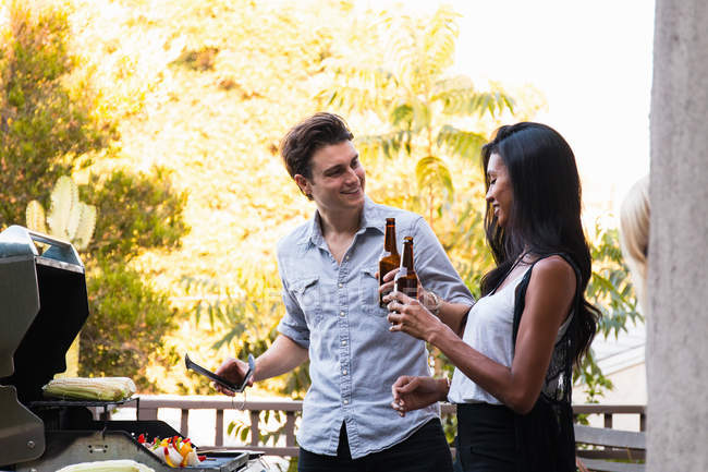 Jeune couple à l'extérieur, tenant des bouteilles de bière, cuisinant des aliments sur le barbecue — Photo de stock