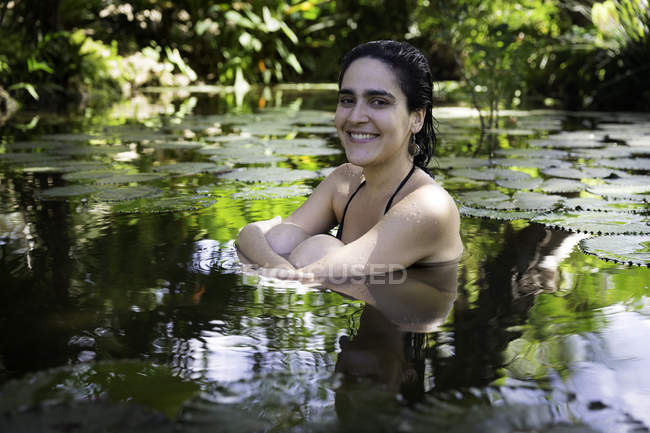 Mujer joven rodeada de almohadillas de lirio sentada en el estanque abrazando las rodillas, mirando a la cámara sonriendo - foto de stock