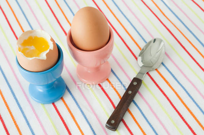 Boiled eggs in egg stands with spoon on striped tableloth — Stock Photo