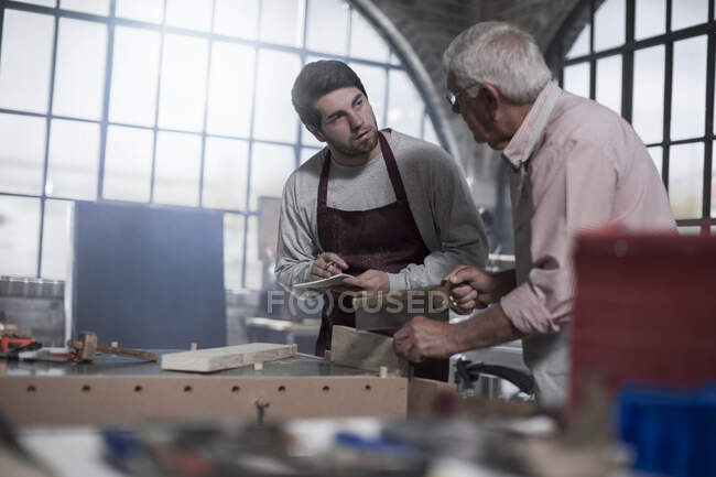 Кейптаун, Южная Африка, молодой деревообработчик записывает заметки от пожилого человека в мастерской — стоковое фото