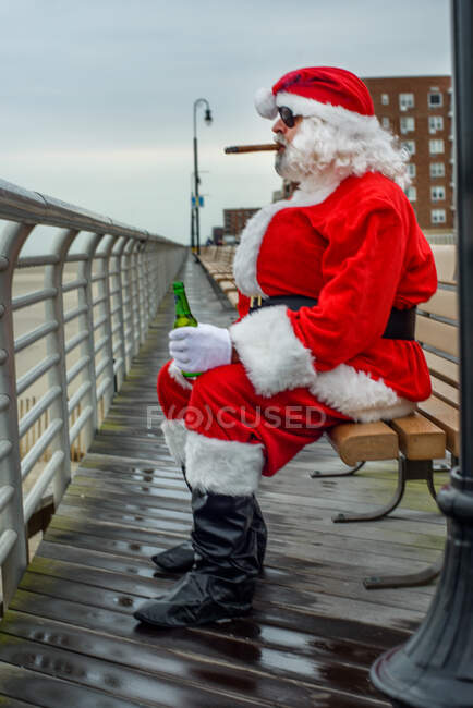 Man dressed in Santa suit, sitting on bench, smoking cigar, holding bottle of beer — Stock Photo