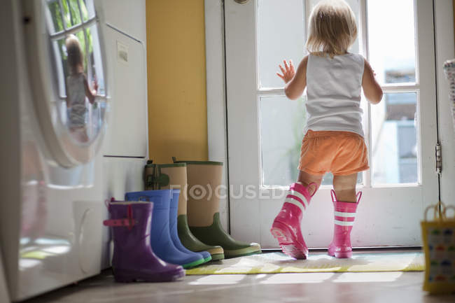 Female toddler wearing rubber boots looking out of back door window — Stock Photo