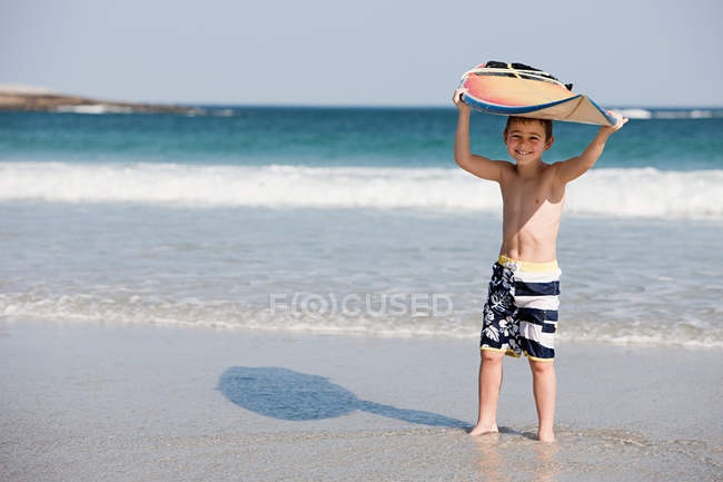 Young boy holding surfboard above head at the edge of the sea — Stock Photo