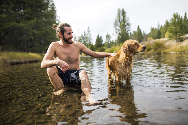 Young man sitting in river petting dog, Lake Tahoe, Nevada, USA — Stock Photo