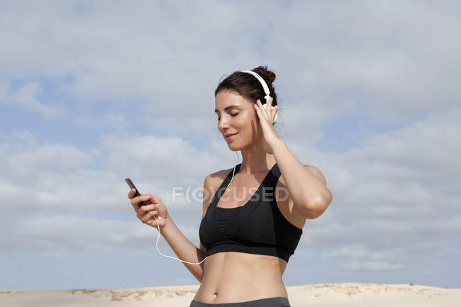 Mujer adulta seleccionando música para auriculares mientras hace ejercicio en la playa - foto de stock
