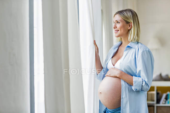 Embarazo a término mujer joven mirando por la ventana - foto de stock