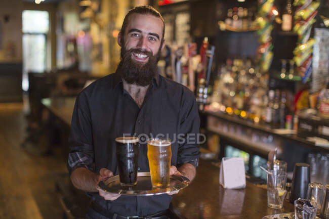 Portrait of young male bartender carrying tray of beer in public house — Stock Photo