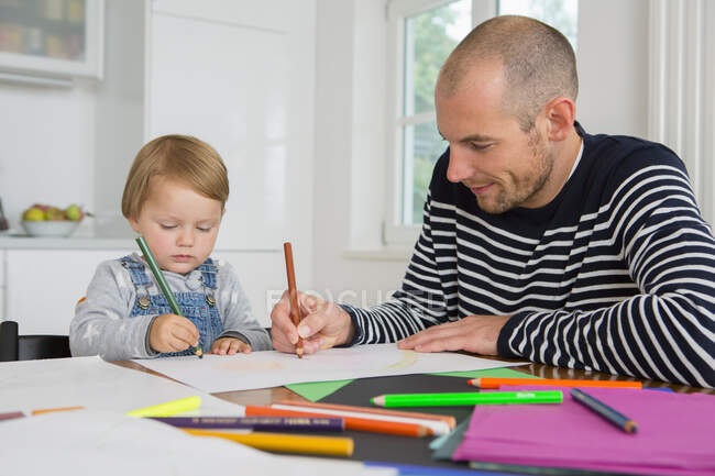 Hombre adulto medio y niña pequeña dibujando en la mesa de la cocina - foto de stock