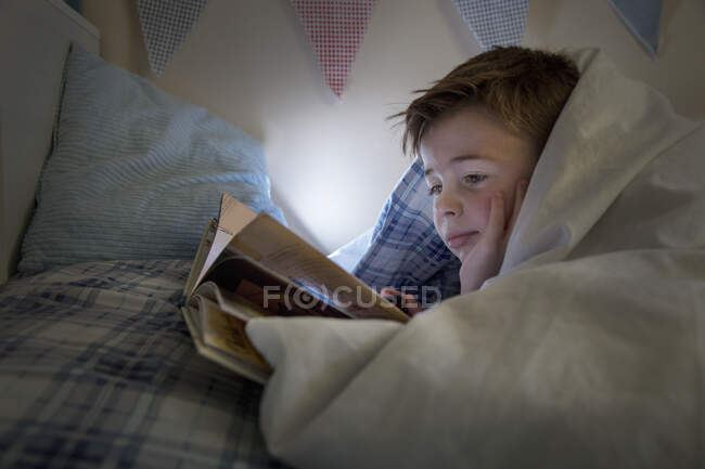 Menino deitado na cama envolto em edredão leitura livro — Fotografia de Stock