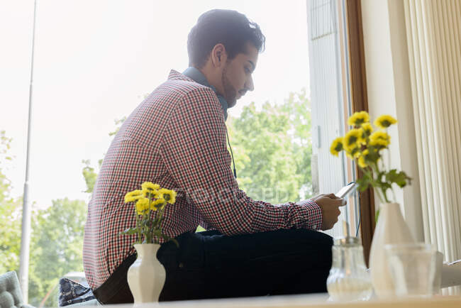 Jeune homme dans un siège de fenêtre de café en sélectionnant la musique sur smartphone — Photo de stock