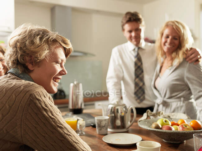 Hijo adolescente sentado en la mesa de la cocina con los padres en el fondo - foto de stock