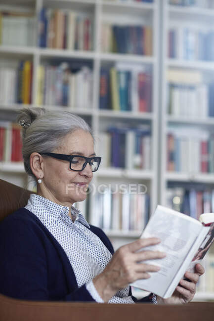 Grey haired mature woman reading book from bookshelves — Stock Photo